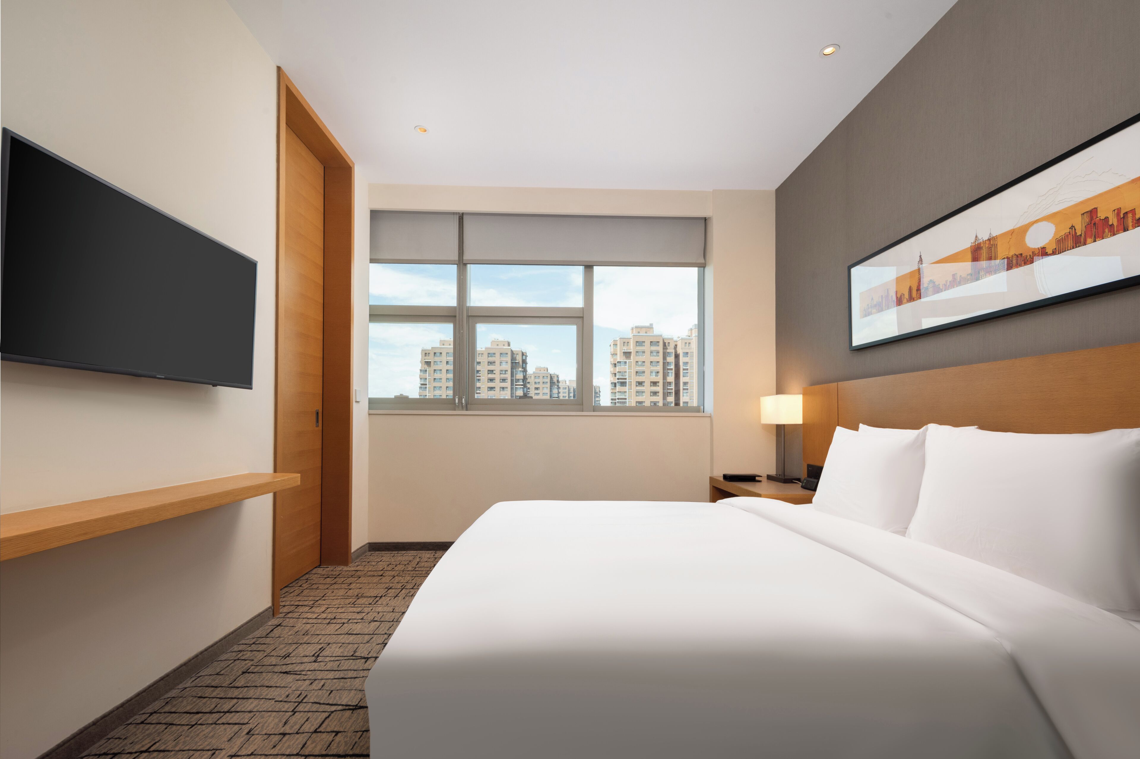 Habitación de hotel con una cama, un televisor montado en la pared, una ventana con vistas a la ciudad y un cuadro enmarcado en la pared.