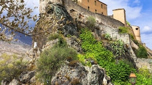 Un edificio de piedra histórico con un muro fortificado, situado en una ladera rocosa con vegetación.