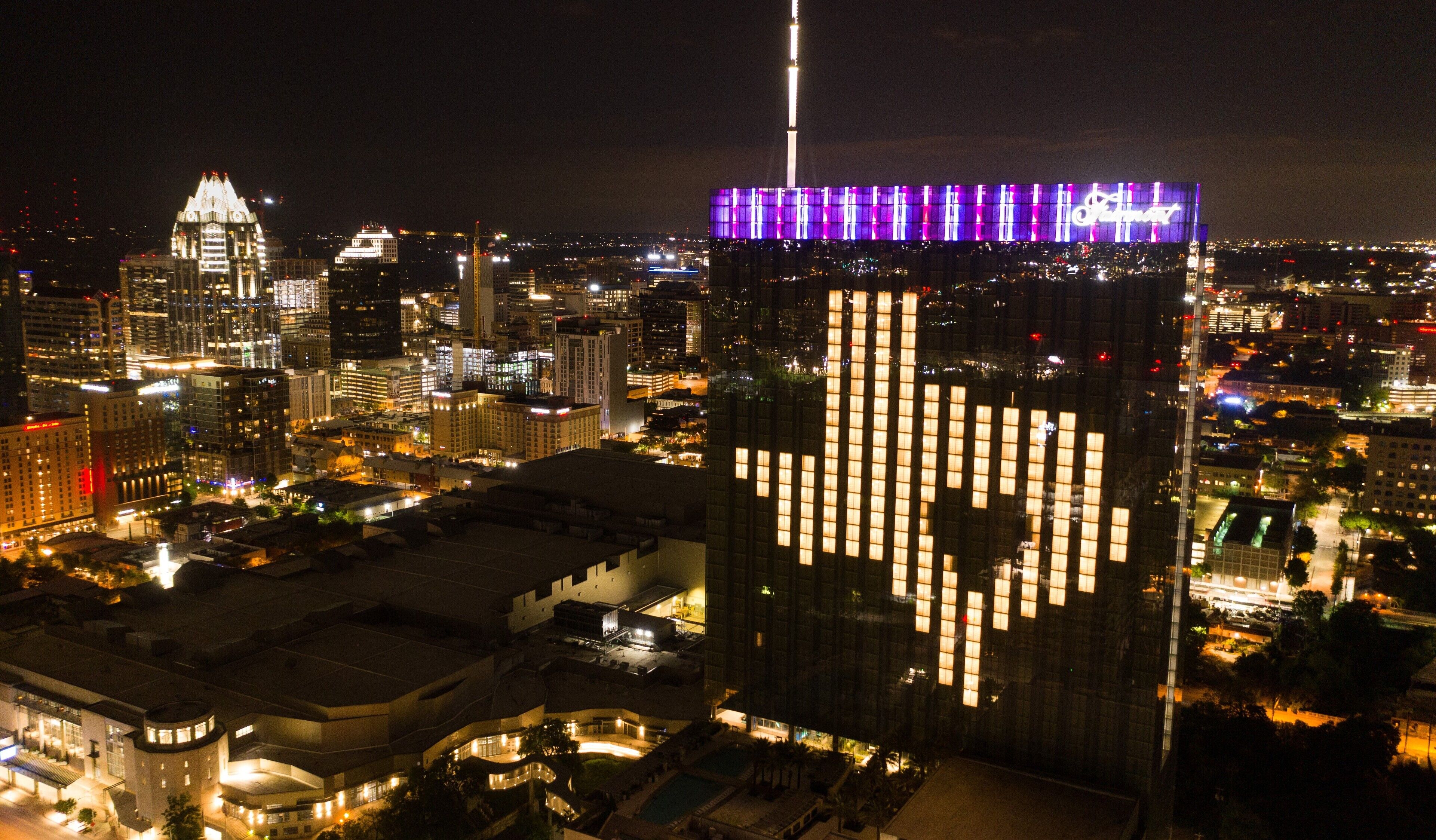 Um edifício alto com um padrão iluminado característico na fachada, cercado por outros edifícios e luzes da cidade à noite.