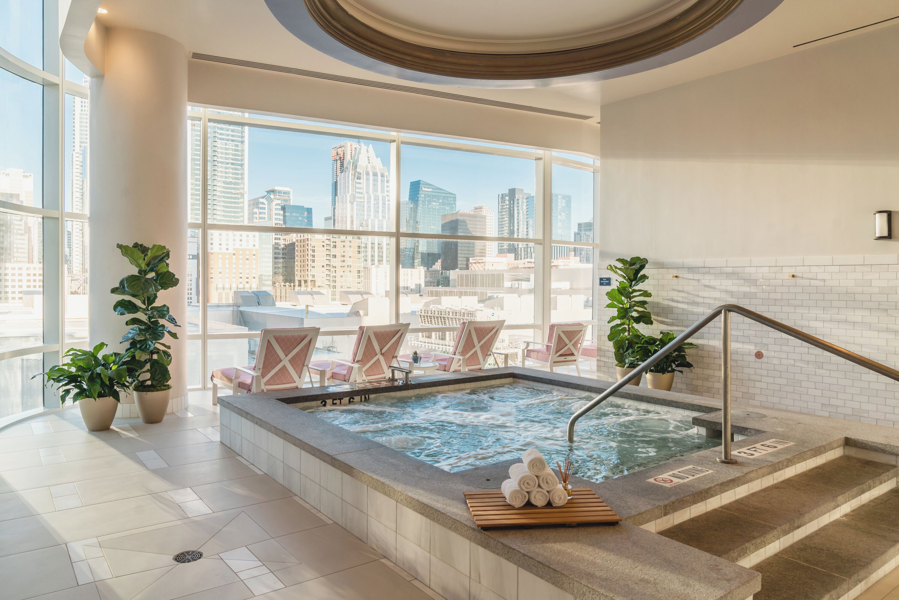 Piscina interior moderna con vistas a la ciudad, rodeada de sillones de descanso y plantas en macetas.
