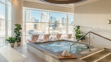 Piscina interior moderna con vistas a la ciudad, rodeada de sillones de descanso y plantas en macetas.