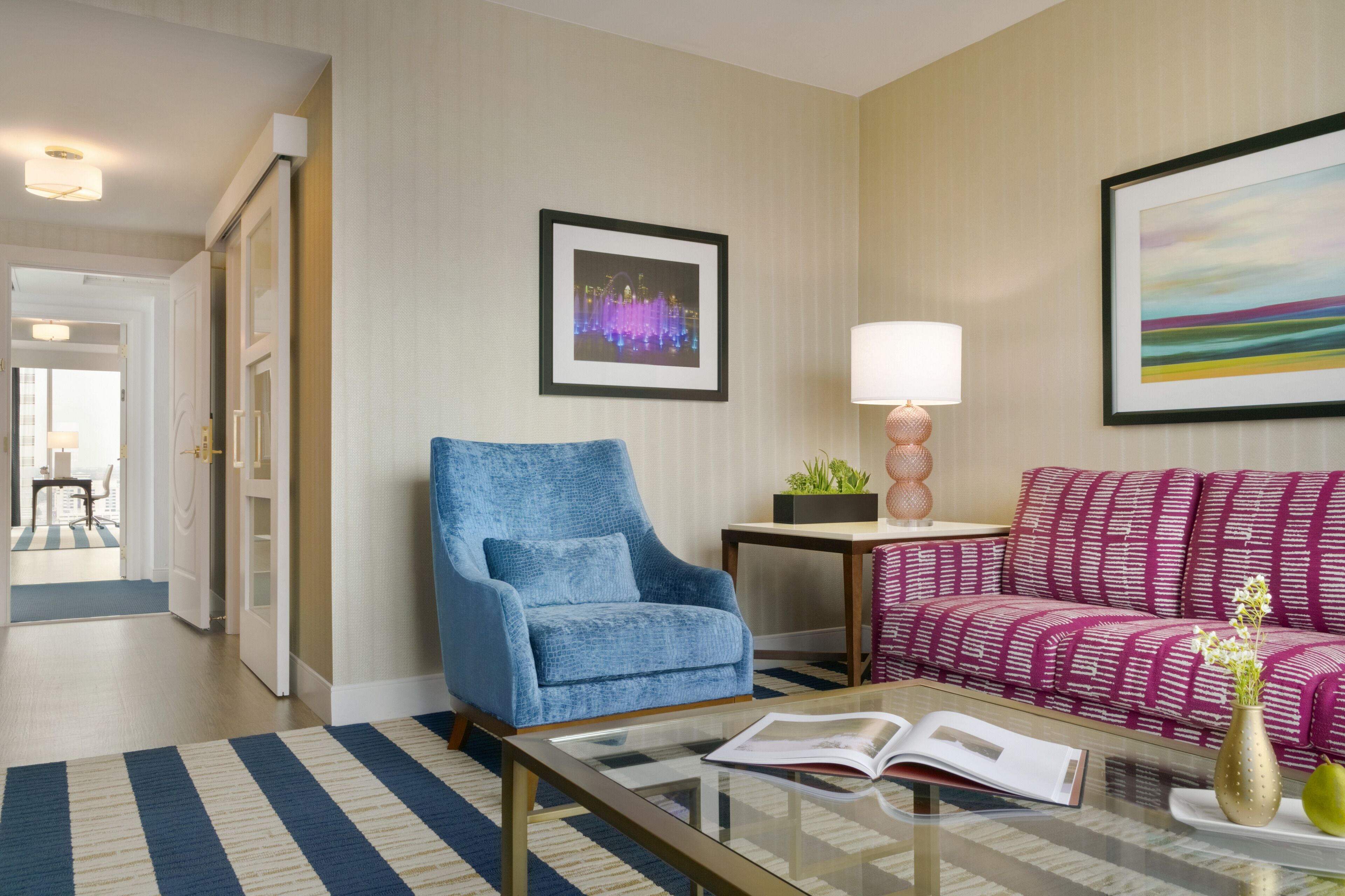 A living room with a blue armchair, a pink patterned sofa, a glass coffee table with a book, a small table with a lamp, and a framed picture on the wall.