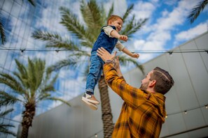 Eine Person hält ein Kind im Freien, im Hintergrund Palmen und ein modernes Gebäude.
