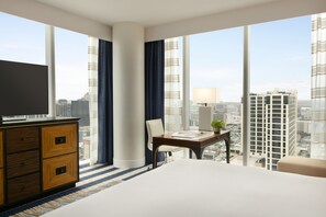Ein Hotelzimmer mit einem großen Bett, einem Schreibtisch, einem Sessel und einem Fernseher. Das Zimmer verfügt über große Fenster mit Blick auf die Stadt.