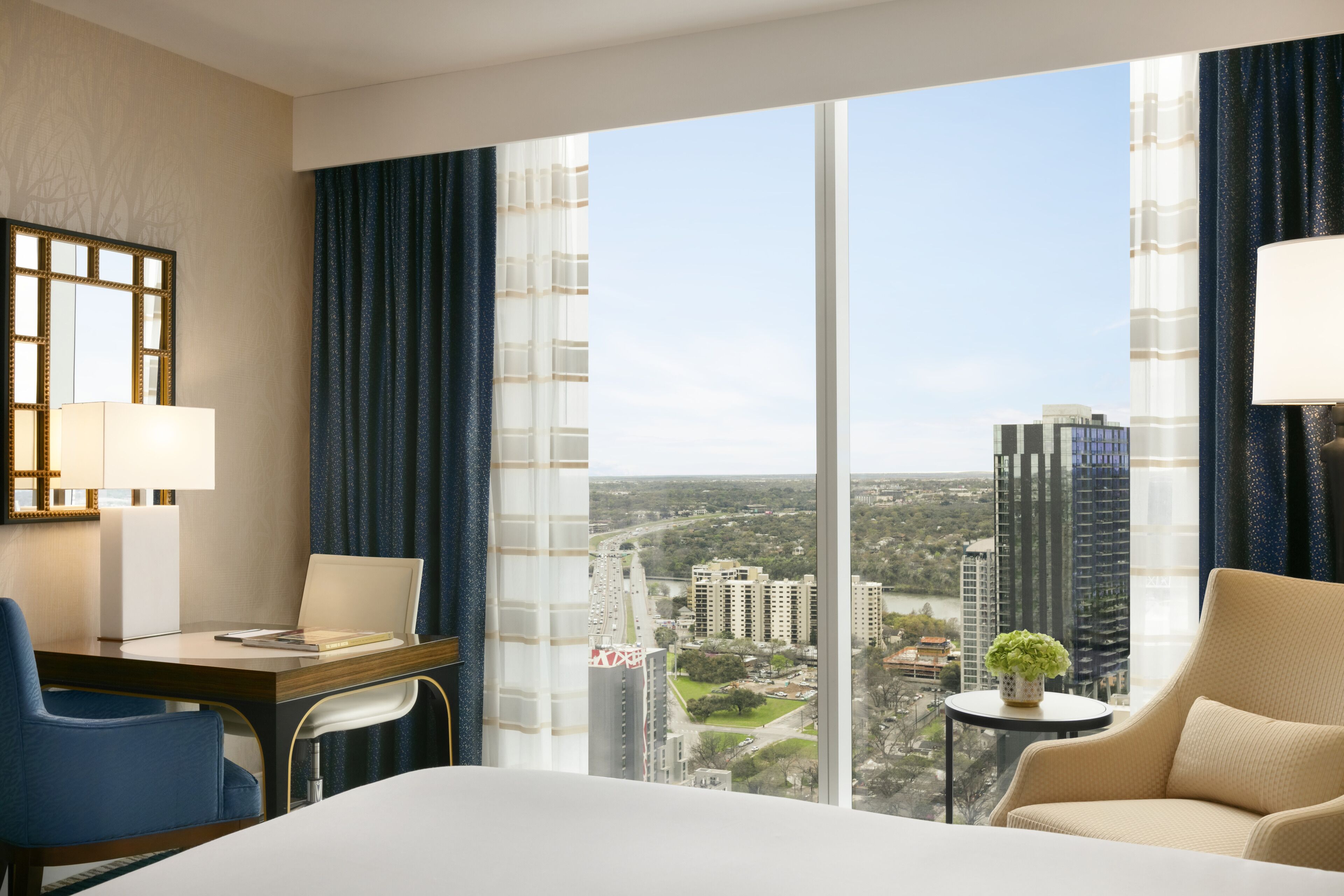 A hotel room with a large window offering a city view, a bed, a desk with a chair, and a small table with a plant.