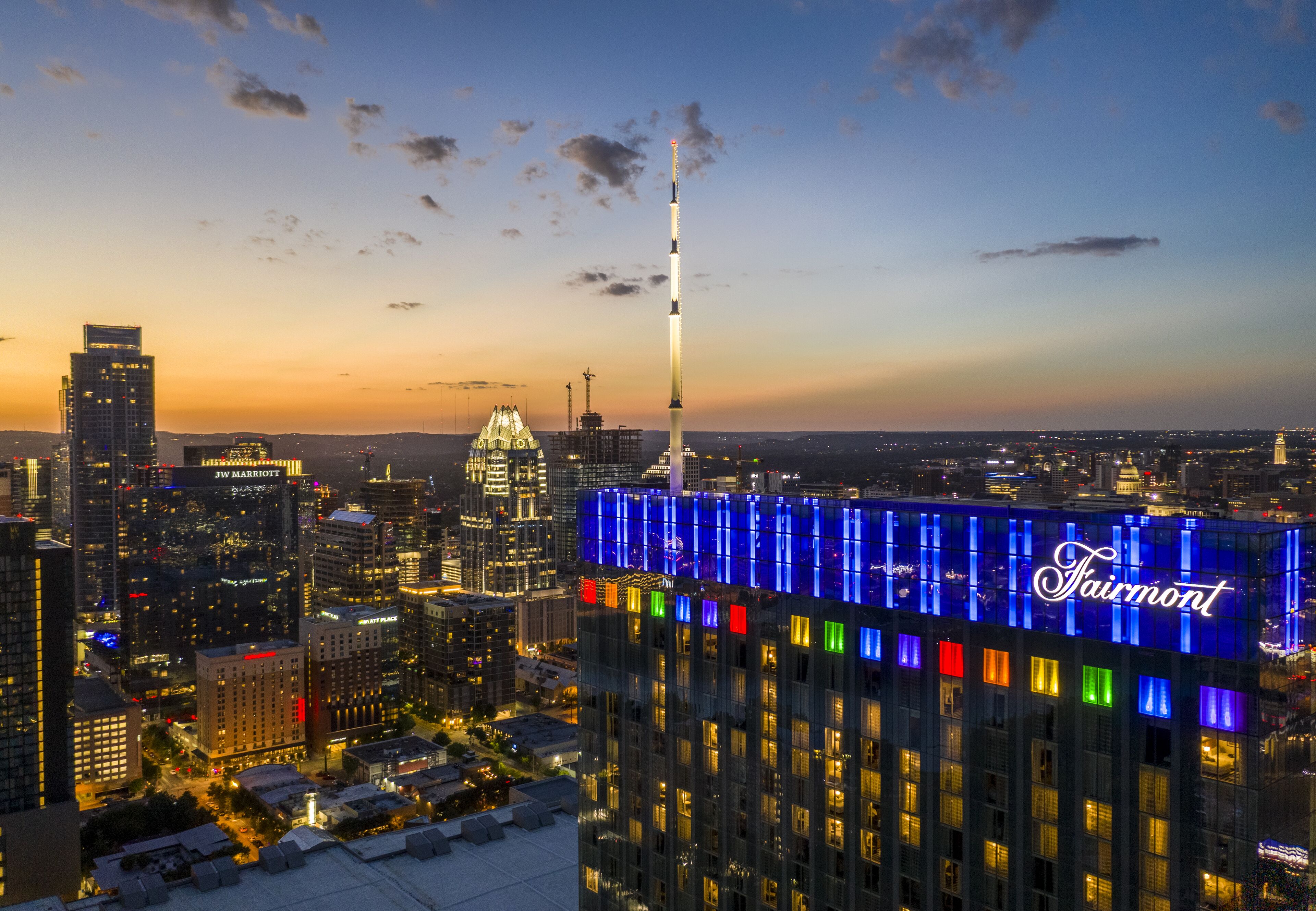 A cityscape at dusk with a prominent building featuring the Fairmont name illuminated in blue.