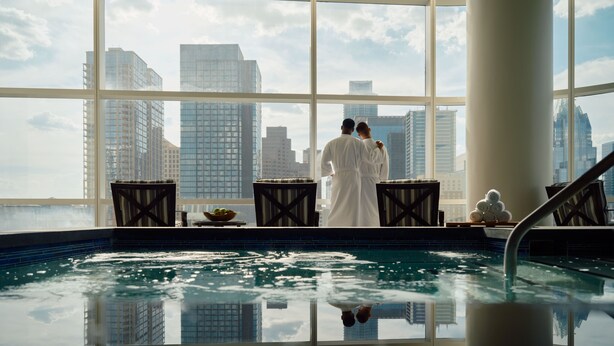 Un couple, vêtu de longues robes blanches, se tient près d’une piscine, avec une vue sur la ville qui s’étend au loin à travers de grandes fenêtres.
