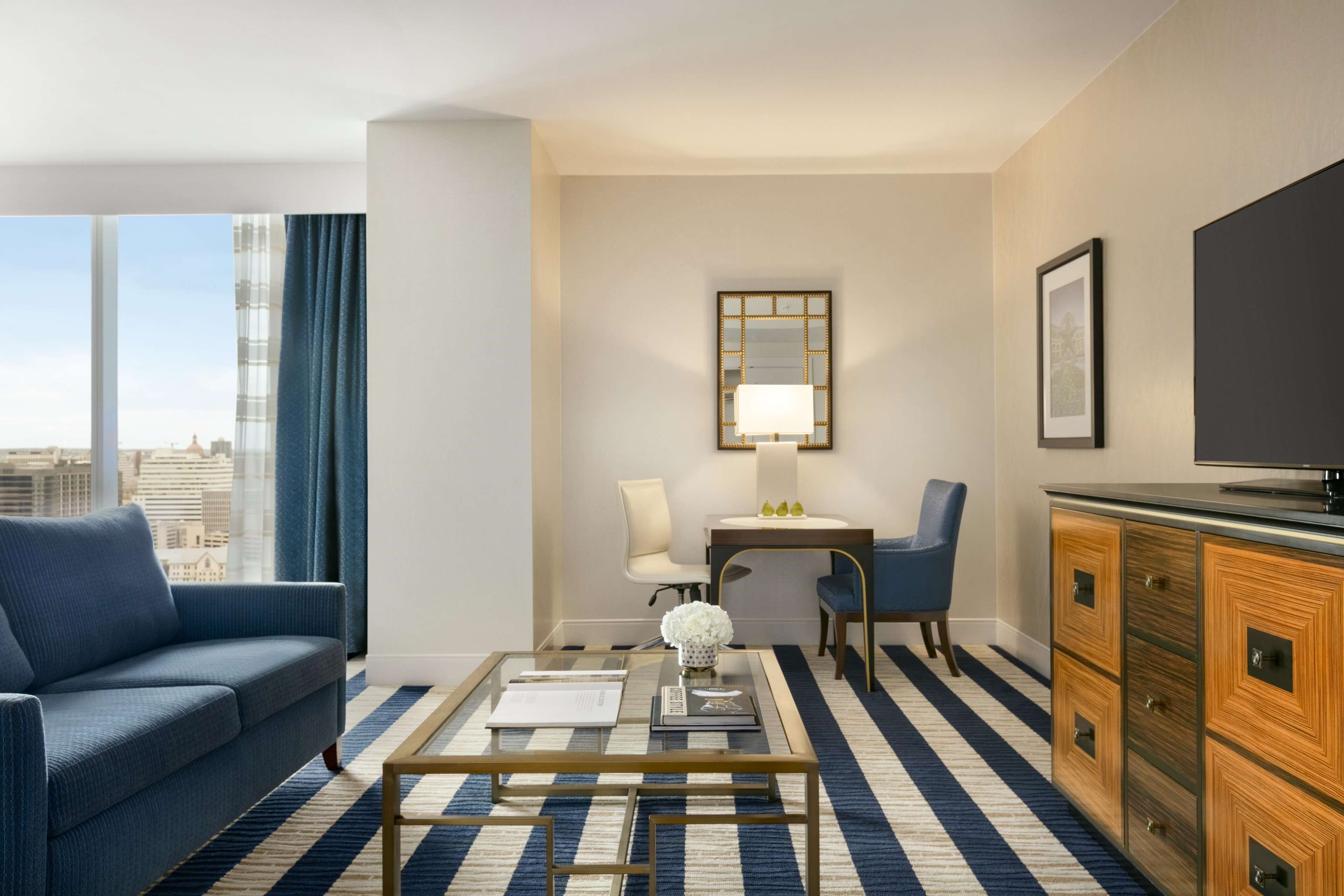 A modern hotel room with a large window, a sofa, a coffee table, a desk with a lamp, a television, and a dresser.