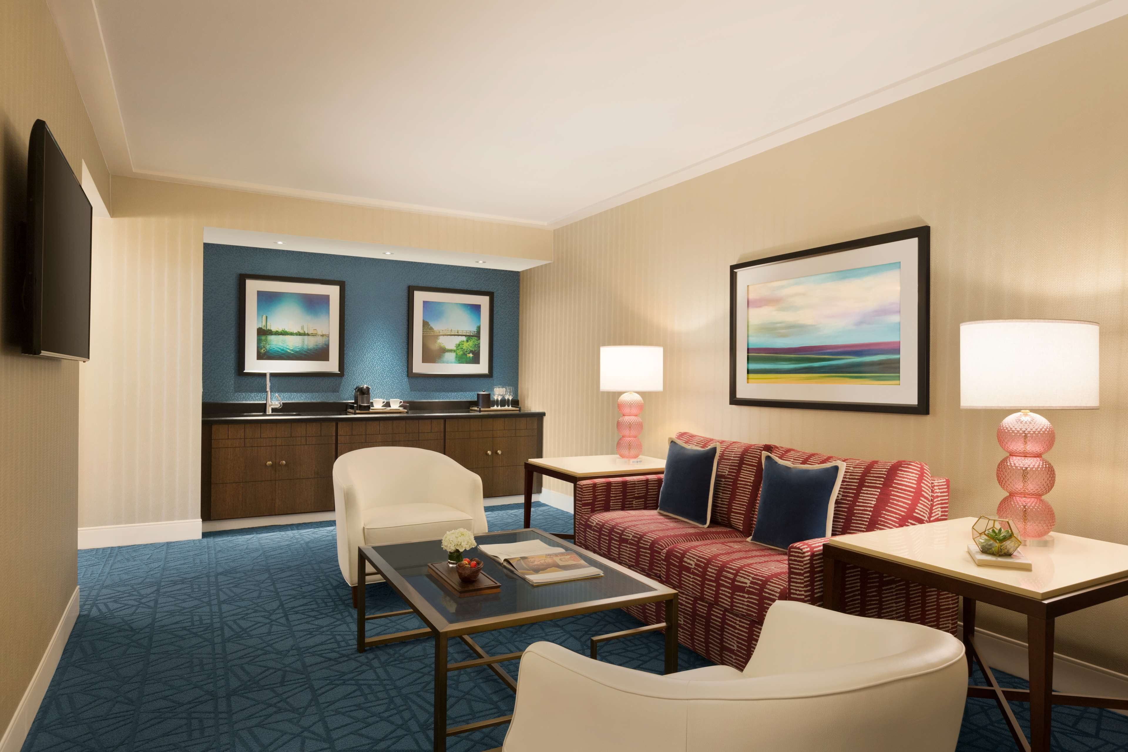 A hotel room with a blue carpet, a white sofa, a red patterned sofa, a white armchair, a small table with a lamp, a TV mounted on the wall, and framed pictures on the wall.
