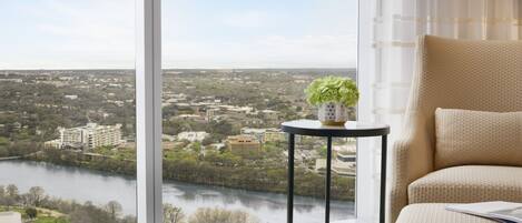 A room with a large window offering a view of a cityscape and a river. There is a round table with a vase of flowers and a chair with cushions.