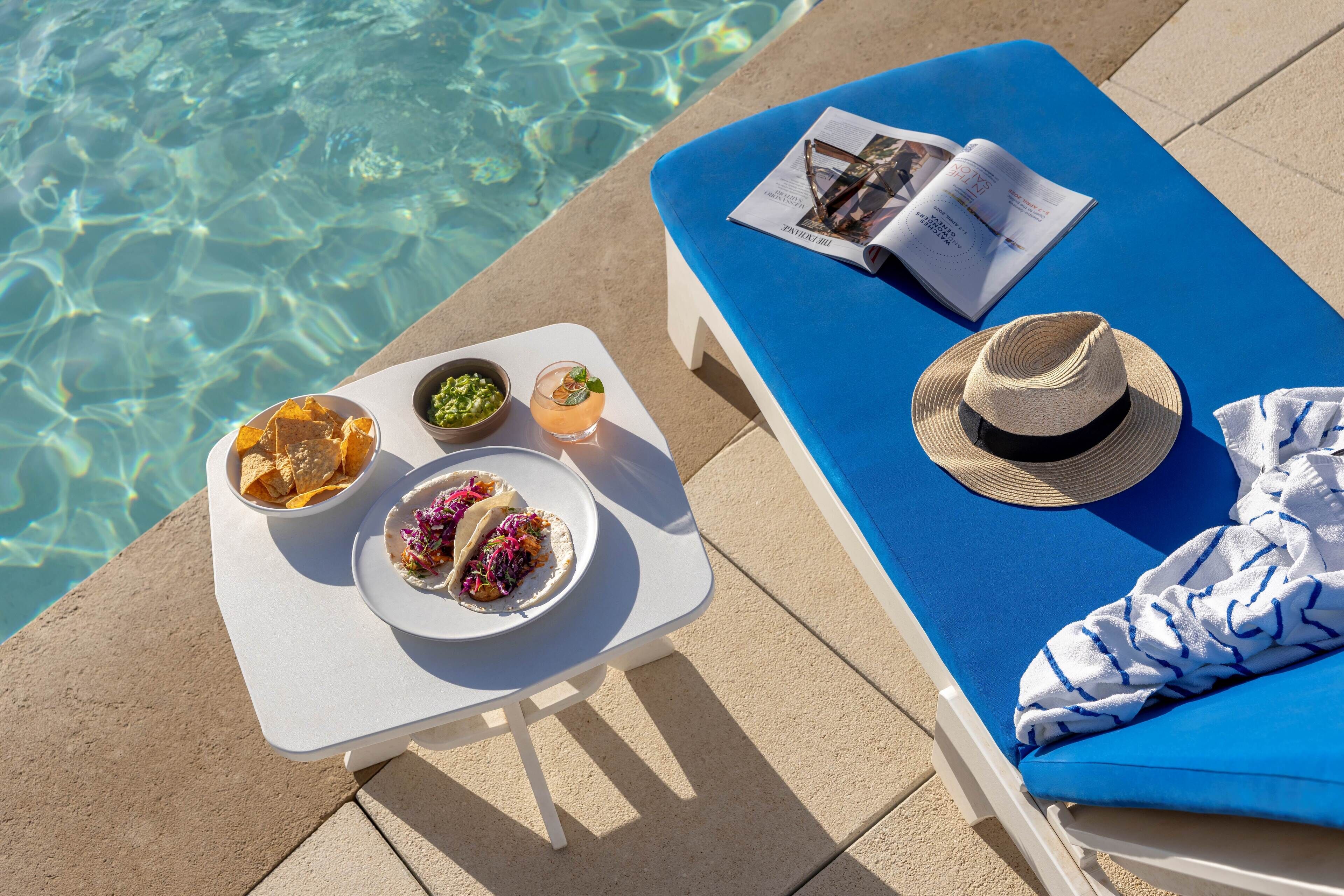 Escena junto a la piscina con una silla de descanso azul, una mesa con comida y bebidas, y una revista.
