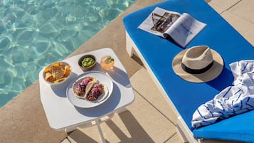 Escena junto a la piscina con una silla de descanso azul, una mesa con comida y bebidas, y una revista.