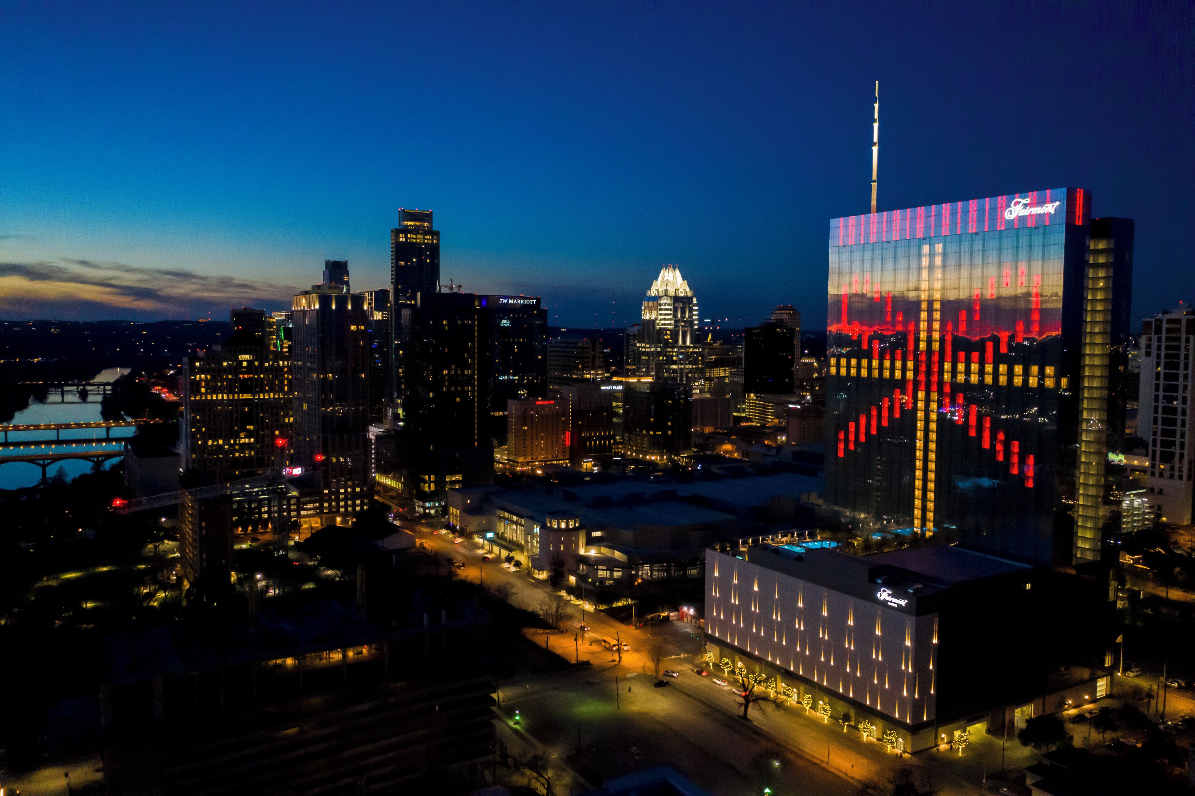 A cityscape at night with illuminated buildings, including a prominent hotel with a distinctive facade and a tall building with a spire.