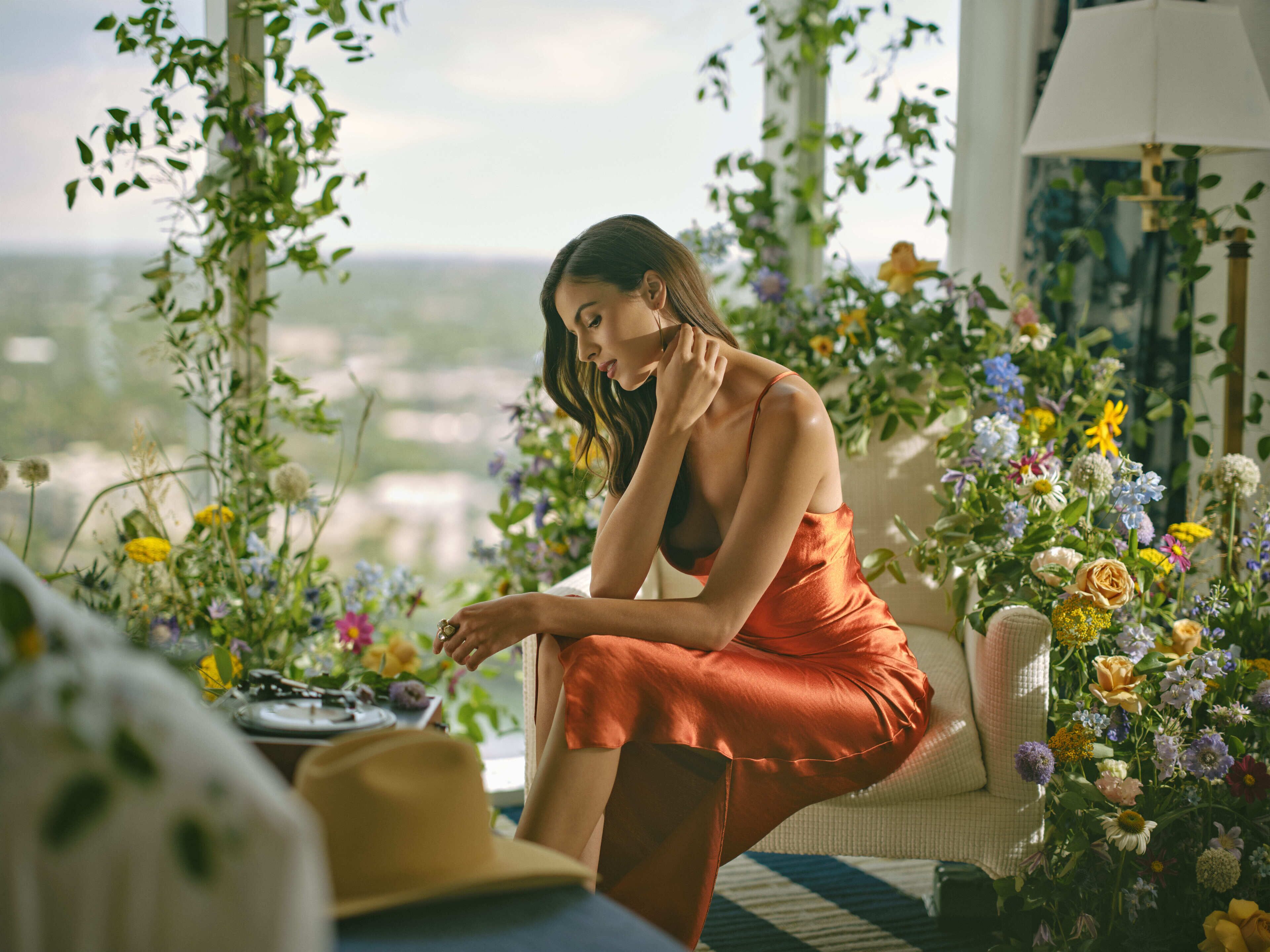 A woman seated in a room with a view of the city, surrounded by various flowers and plants.