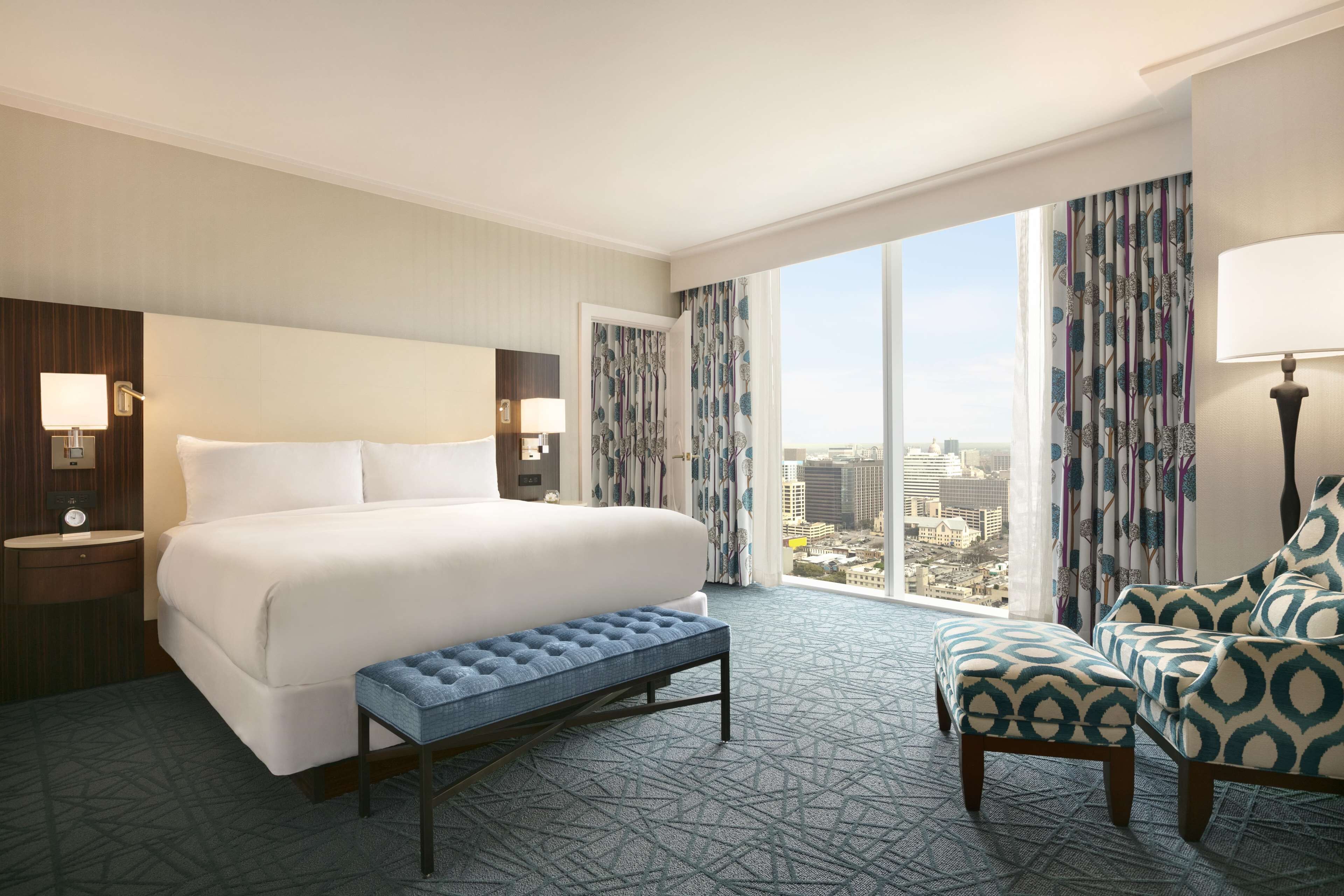 A hotel room with a large bed, a bench, a chair, and a view of the city through large windows.
