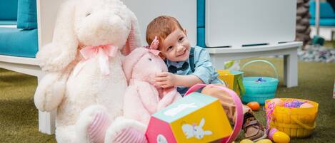 A child sitting on a white bench with a large white stuffed bunny and a smaller pink stuffed bunny. There are colorful toys and a yellow basket on the grass.