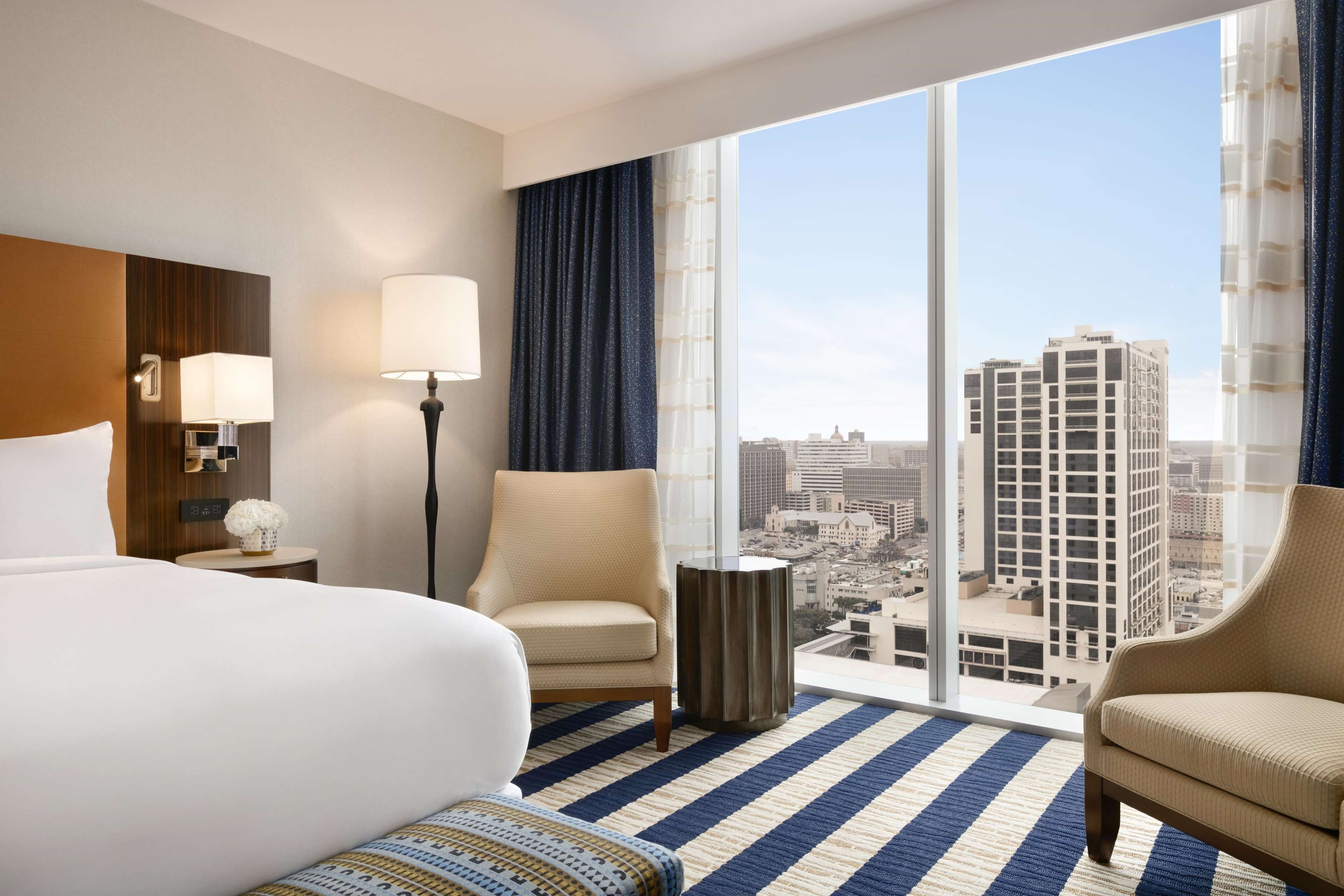 A hotel room with a large bed, two armchairs, a floor lamp, and a view of a cityscape through large windows.