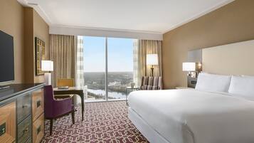 A hotel room with a large bed, a desk with a chair, a TV, and a view of a cityscape through the windows.