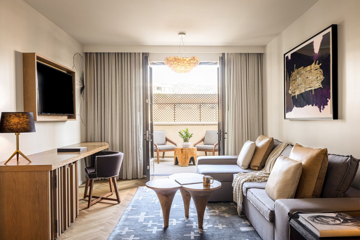 A modern living room with a sofa, coffee table, and chairs. There is a TV mounted on the wall and a window with curtains.