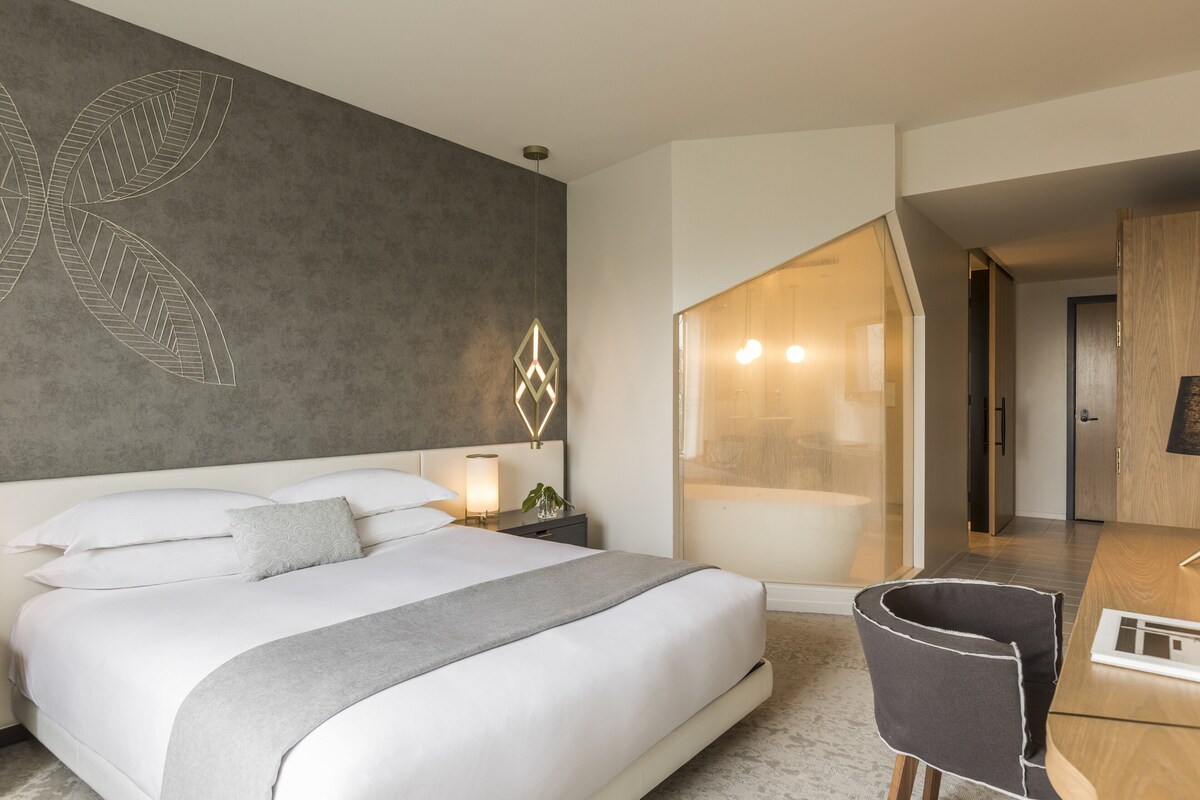 A modern hotel room with a large bed, a desk, and a bathroom visible through an open door.