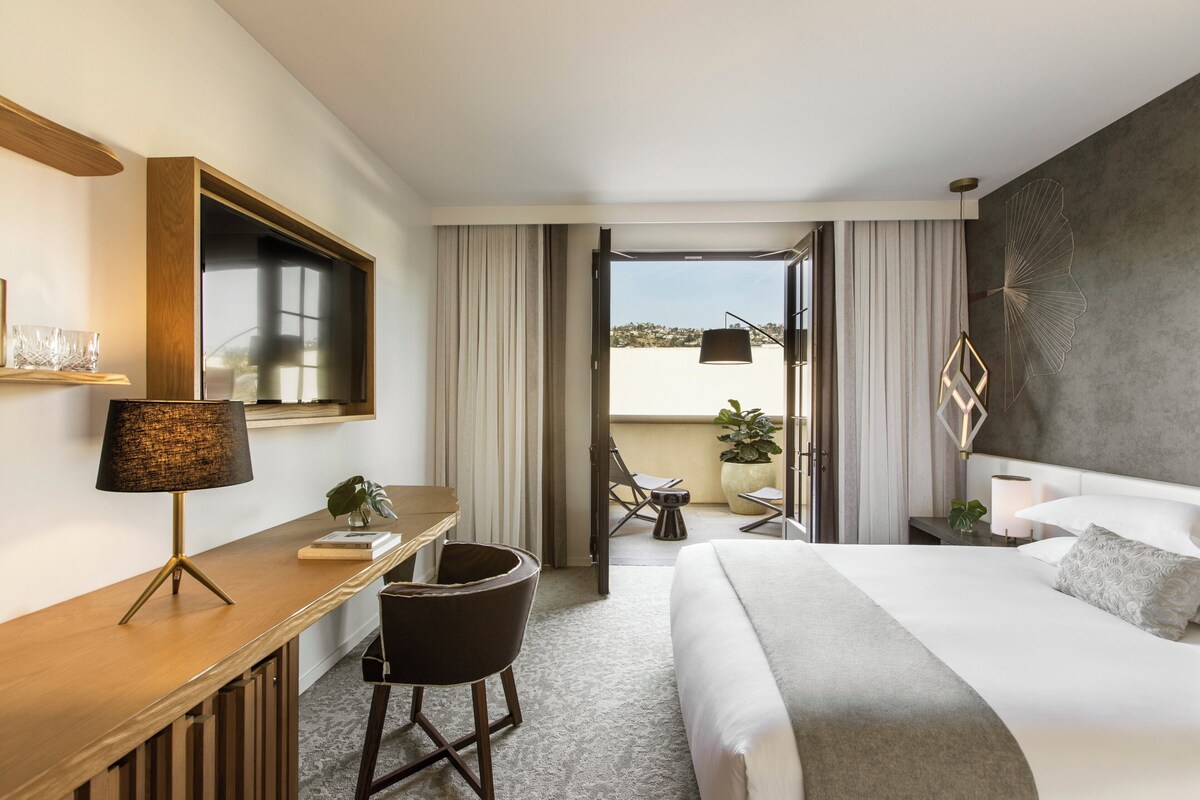 A hotel room with a bed, desk, chair, and a view of the outdoors.