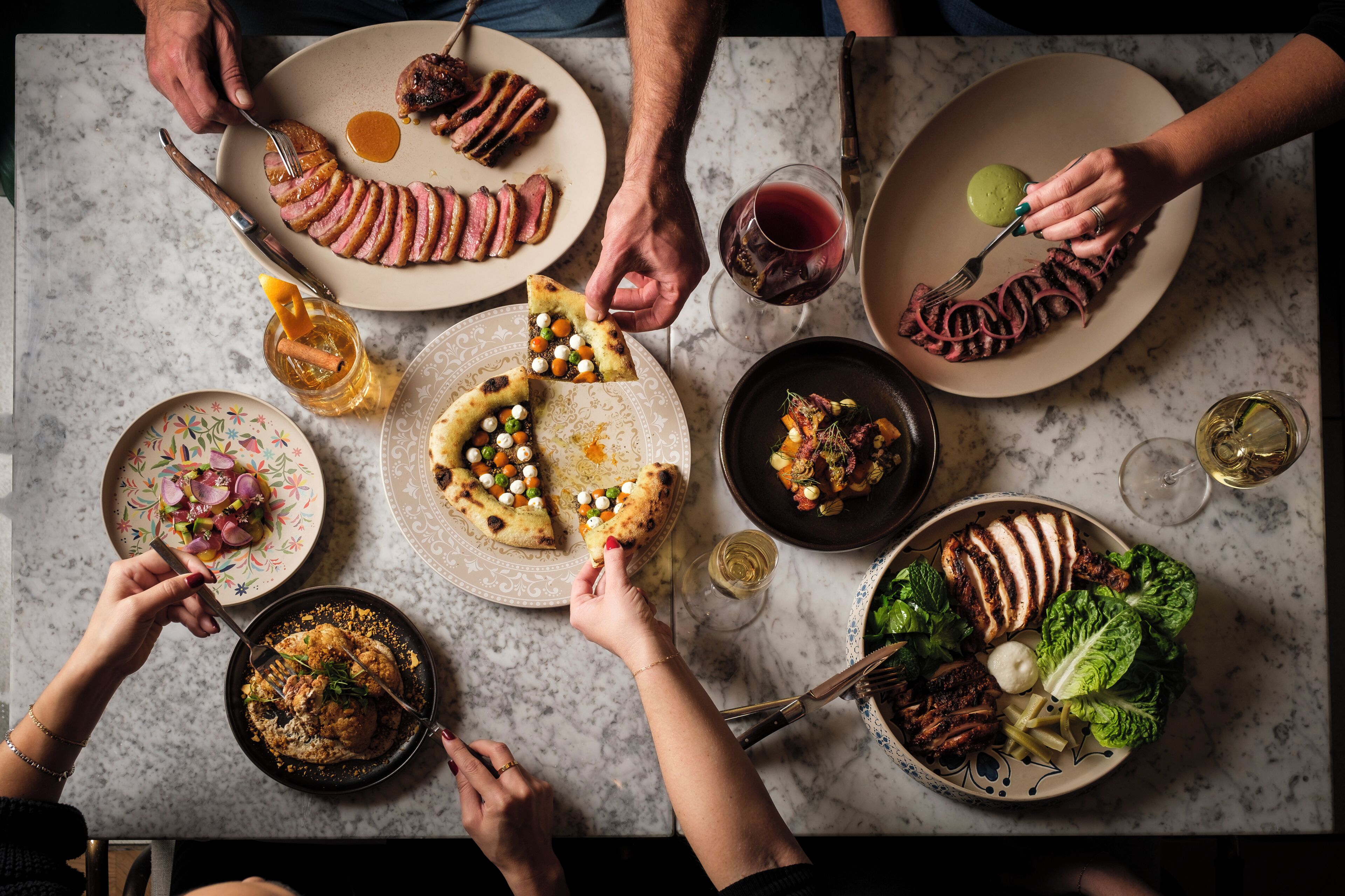 A group of people dining at a table with various dishes, including pizza and meat, and wine glasses.