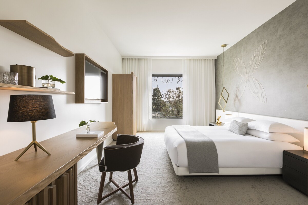 A modern hotel room with a large bed, a desk with a lamp, a chair, a bedside table, and a view of the outdoors through a window.