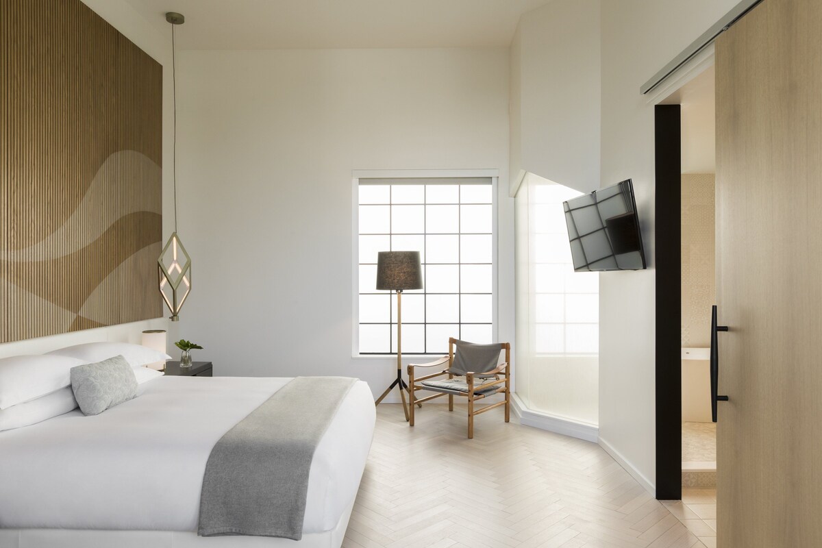 A modern bedroom with a large bed, a desk, a chair, a floor lamp, and a TV mounted on the wall.