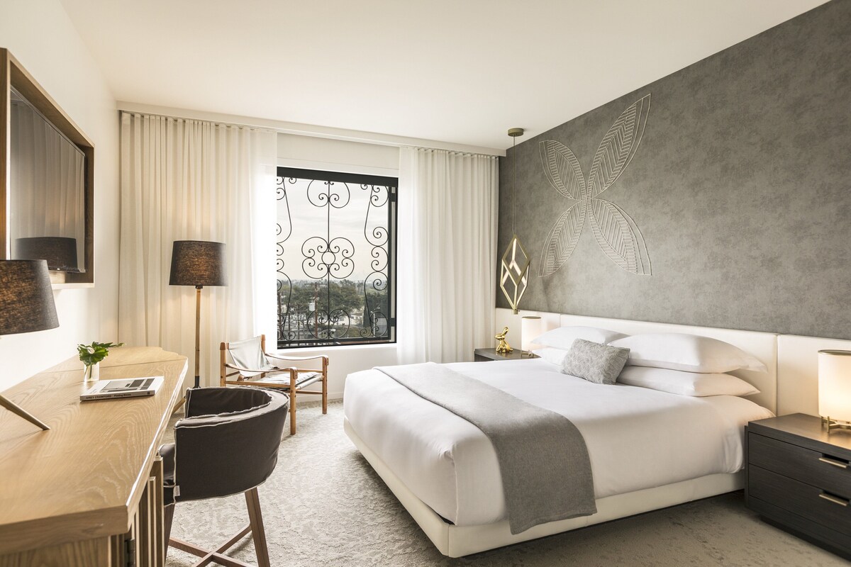 A modern hotel room with a large bed, a desk, a chair, and a view of the outdoors through a window with curtains.