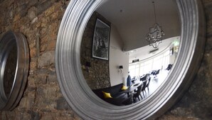 A circular mirror reflecting a room with a stone wall, a black leather sofa, a chandelier, and a dining area with tables and chairs. - The Lawrence (Athboy)