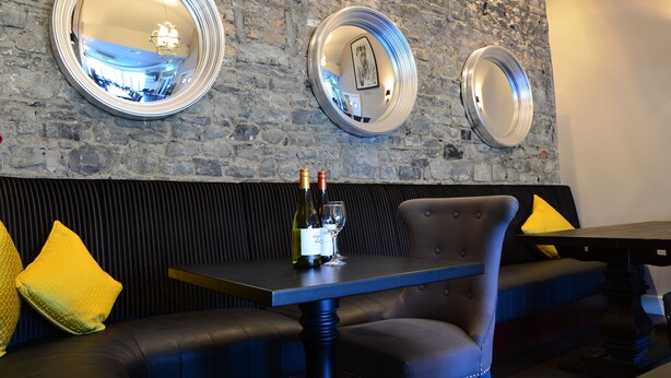 A restaurant interior with a stone wall, three round mirrors, a table with a bottle and glass, and yellow cushions on a bench. - The Lawrence (Athboy)