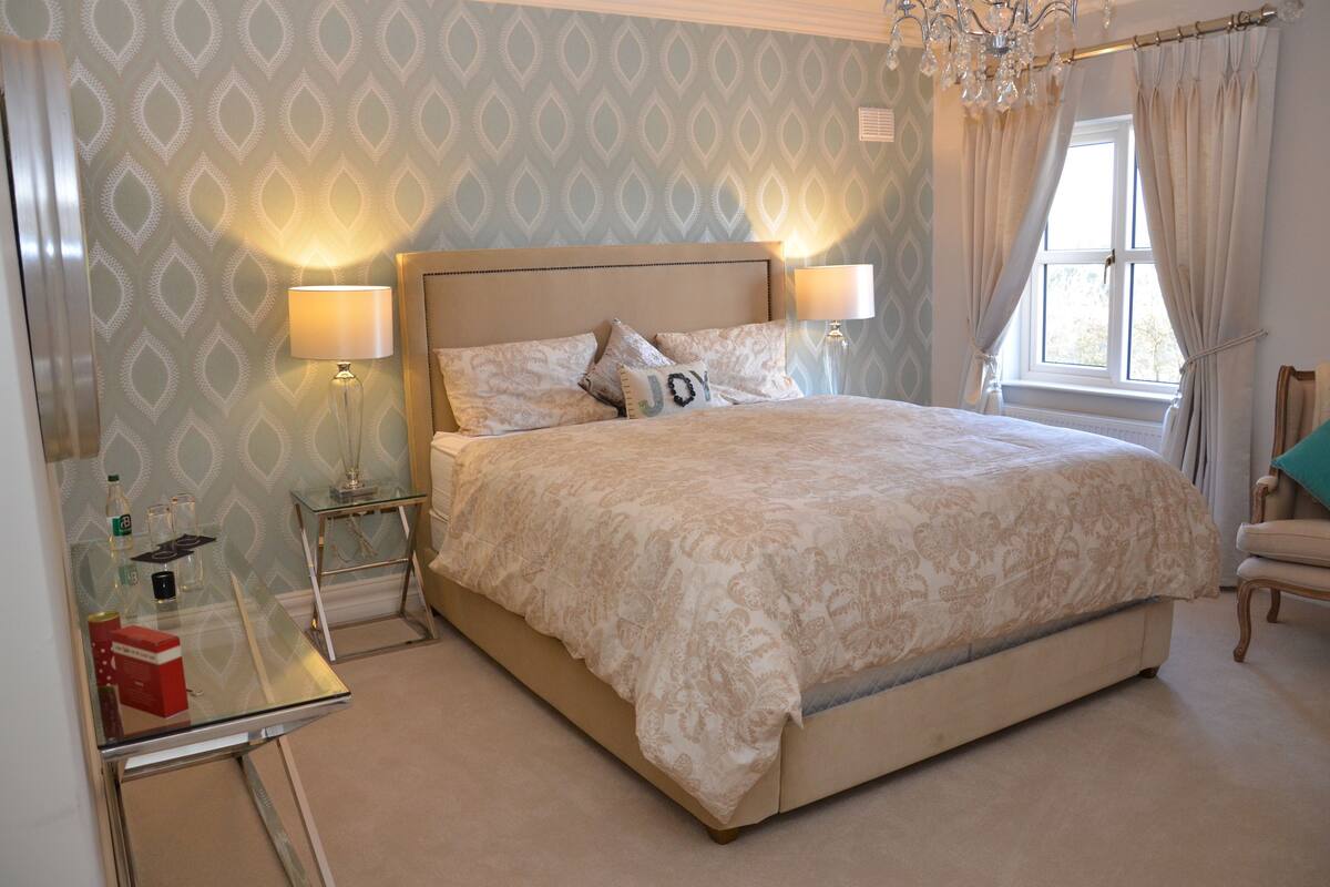 A neatly arranged bedroom with a large bed, bedside lamps, a glass table with a bottle and glasses, and a window with curtains.