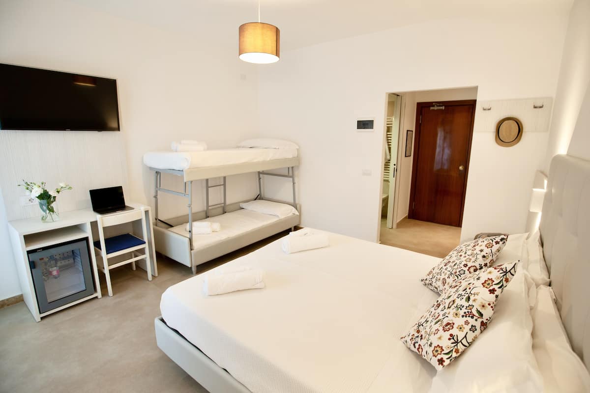 A hotel room with a bunk bed, a desk with a laptop, a small fridge, and a TV mounted on the wall.
