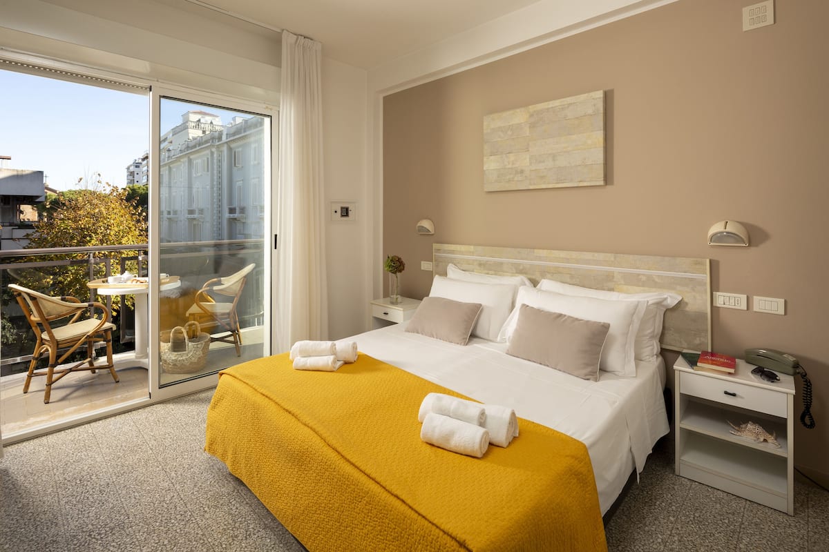 A hotel room with a bed, a nightstand, a chair, a table, and a balcony with a view of buildings.