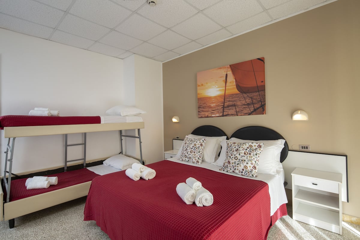 A hotel room with a bunk bed, a single bed with floral pillows, a painting of a sunset, and a nightstand with a lamp.