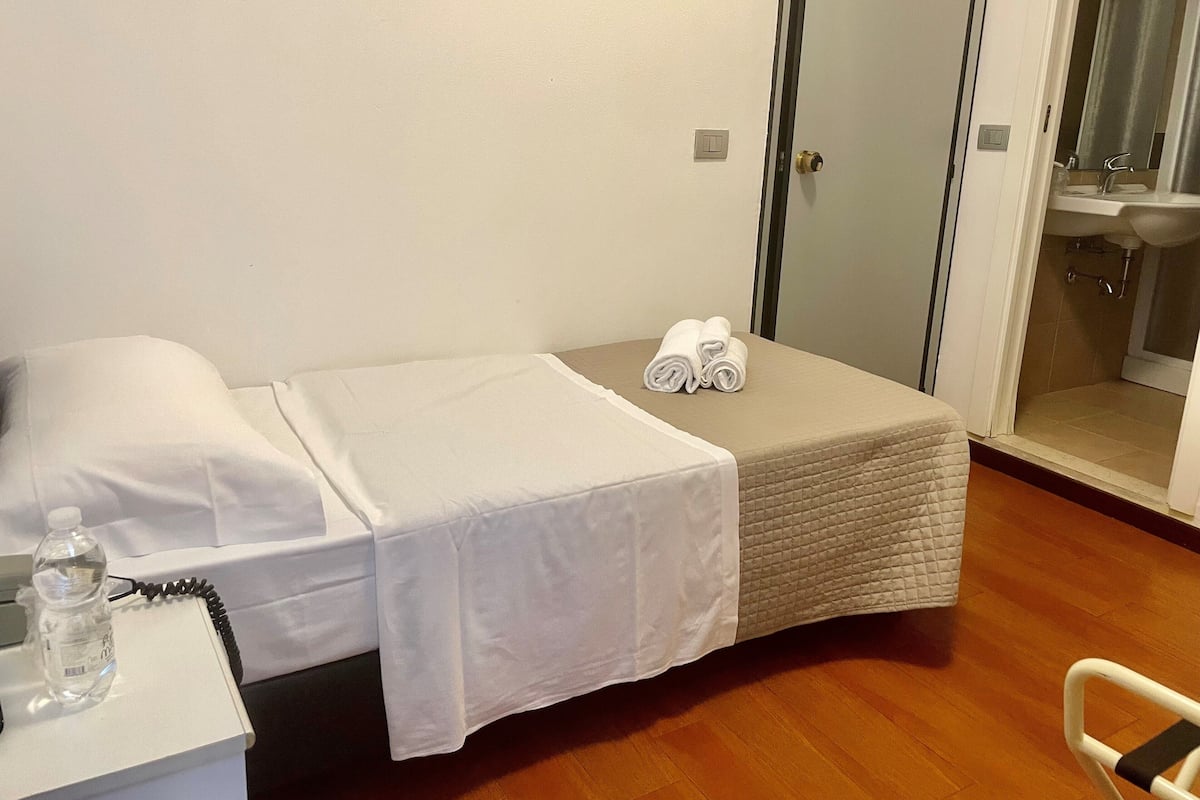 A small, neatly made bed with a white bedspread and two rolled towels, a bedside table with a water bottle, and a bathroom visible through an open door.