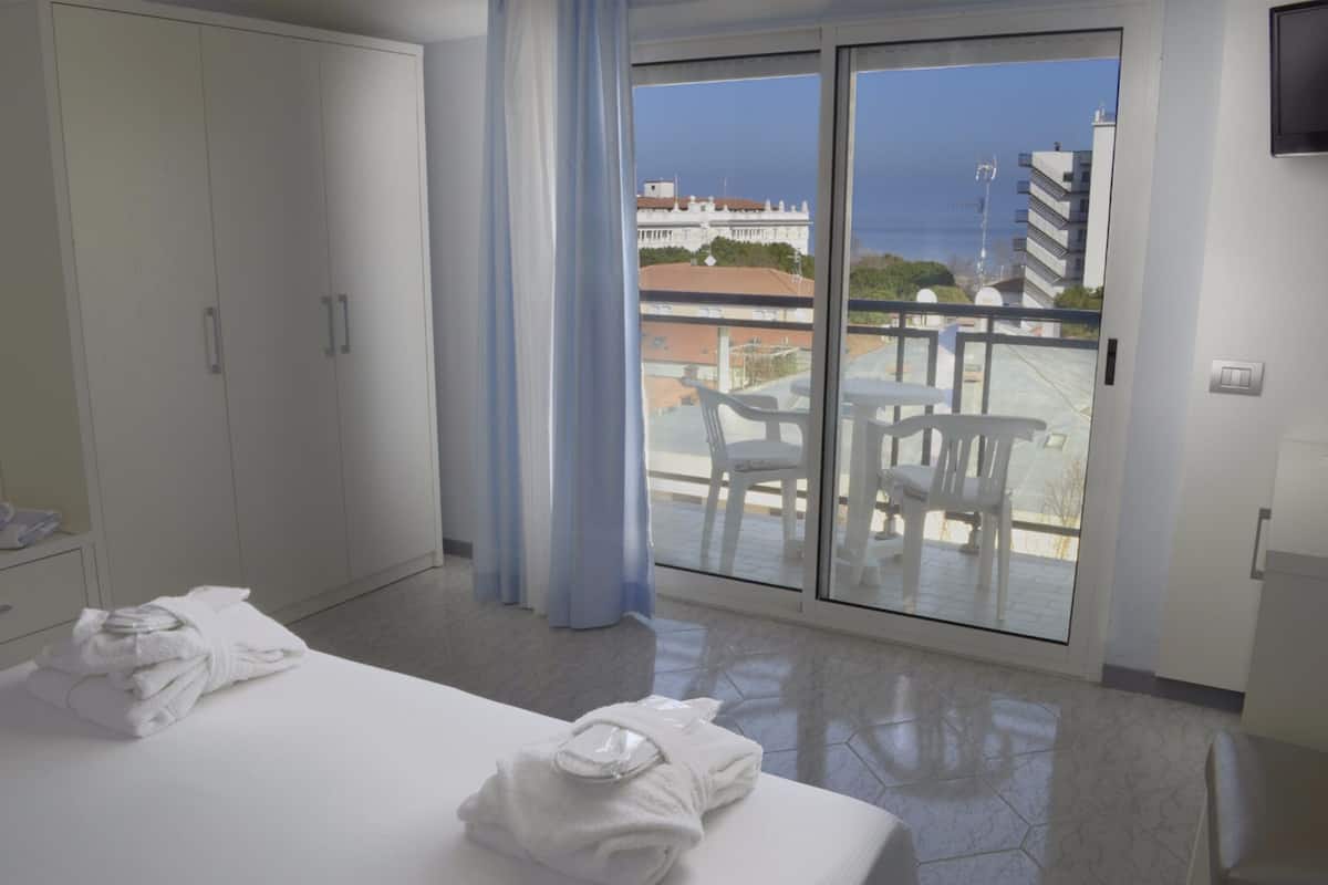 A hotel room with a bed, a set of cabinets, a balcony with a view of the sea and buildings.