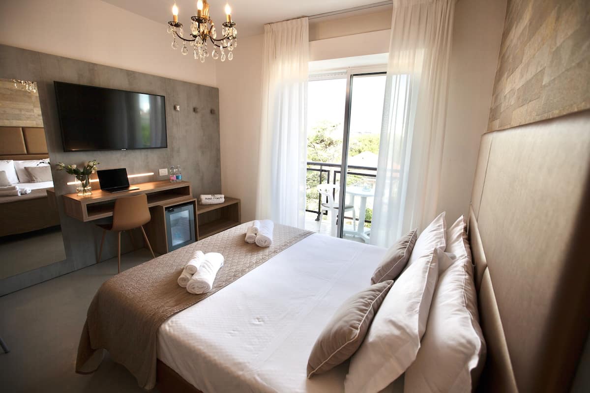 A modern hotel room with a large bed, a desk with a laptop, a flat-screen TV, and a balcony with outdoor furniture.