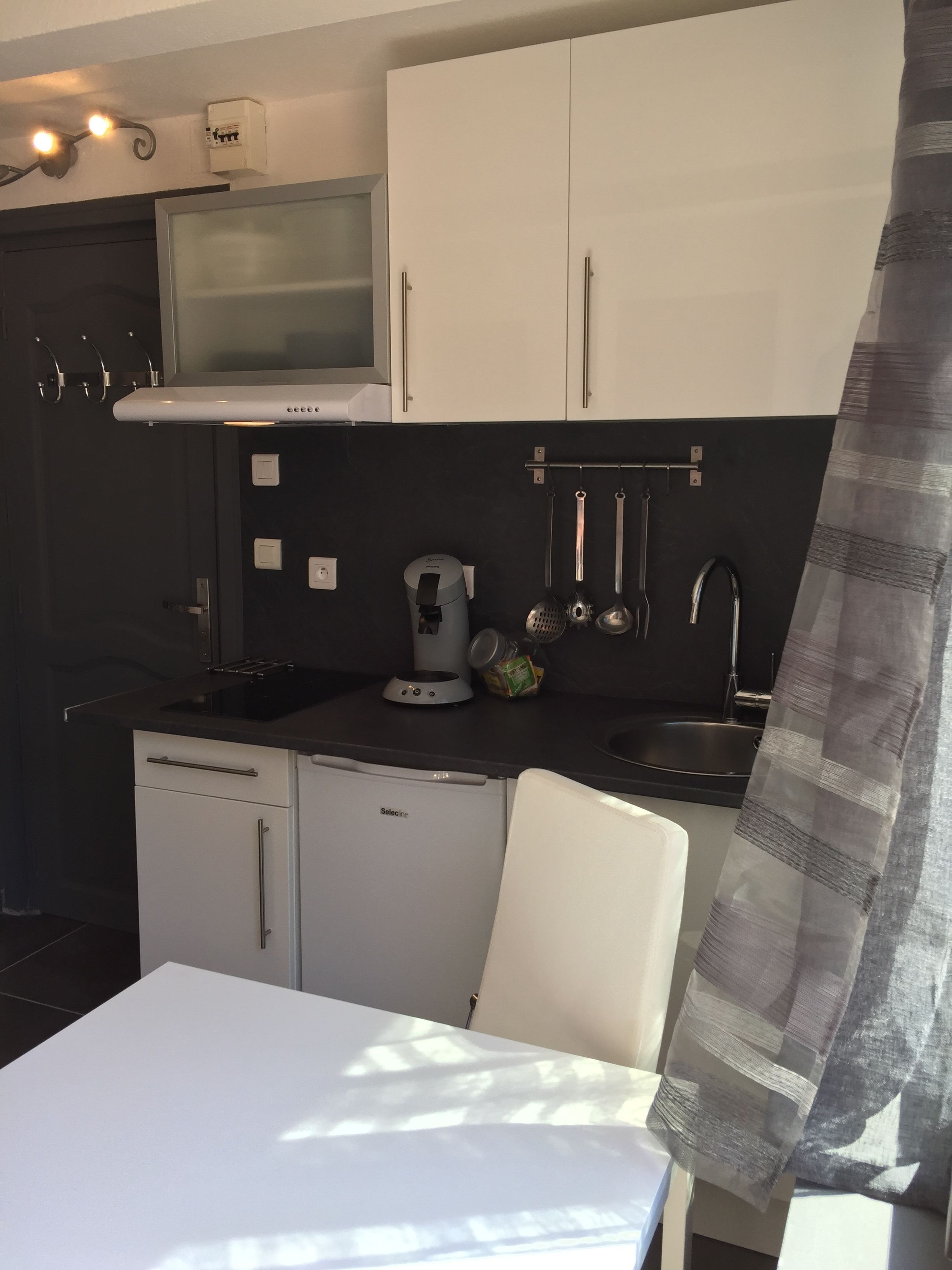 Una cocina compacta con armarios blancos, un refrigerador pequeño, una cafetera y un lavamanos.