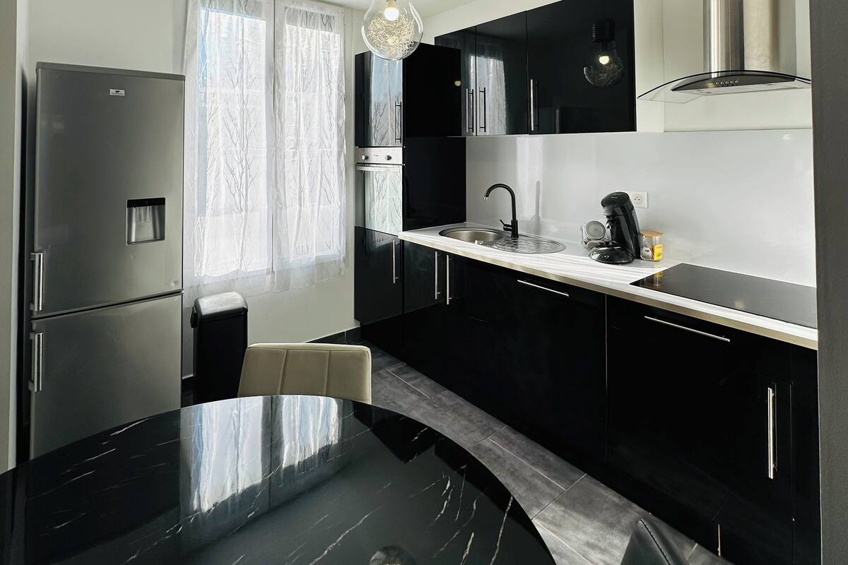 A modern kitchen with black cabinets, a stainless steel refrigerator, a black countertop, and a hanging light fixture.