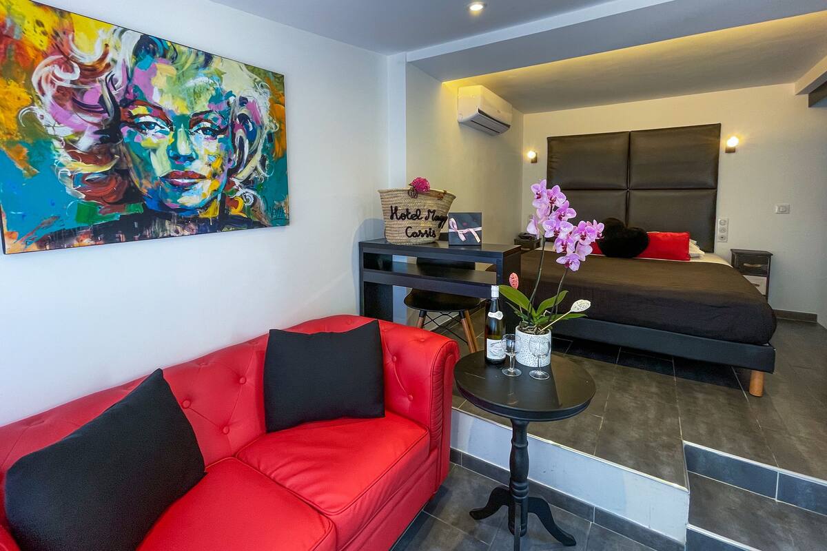 A modern hotel room with a red sofa, a bed, a desk with a computer, a painting on the wall, and a potted orchid.