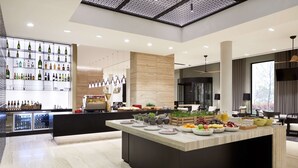 A modern bar area with a variety of bottles and glasses on shelves, a central island with fruits and bowls, and a seating area with a view of the outdoors. - Hyatt Place Melbourne, Essendon Fields (Essendon Fields)