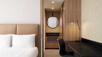 A hotel room with a bed, a desk, a chair, a mirror, and a kitchenette.