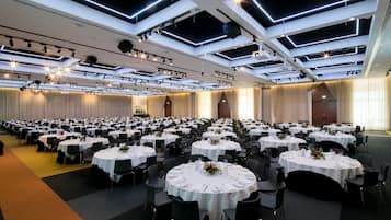 Un amplio salón de banquetes con múltiples mesas redondas, cada una cubierta con manteles blancos y centros de mesa. El techo presenta un diseño moderno con iluminación empotrada y tragaluces.