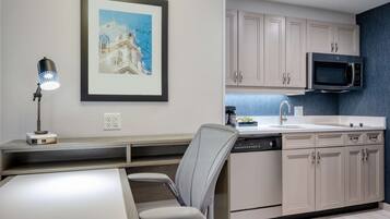 A modern kitchen with a microwave, sink, and cabinets. There is a framed picture on the wall and a desk with a lamp.