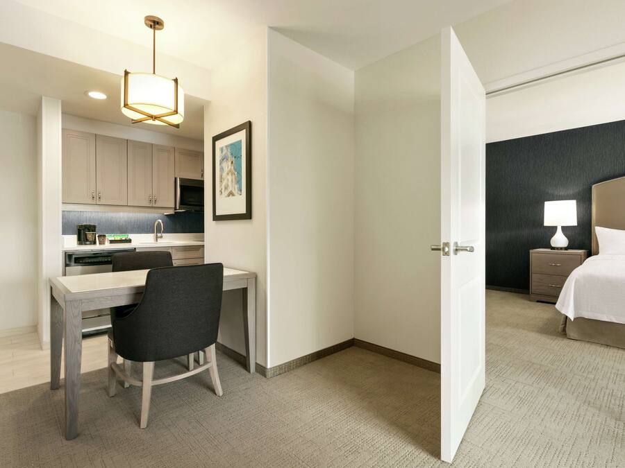 A hotel room with a bed, bedside table, lamp, and a view of the kitchen area.