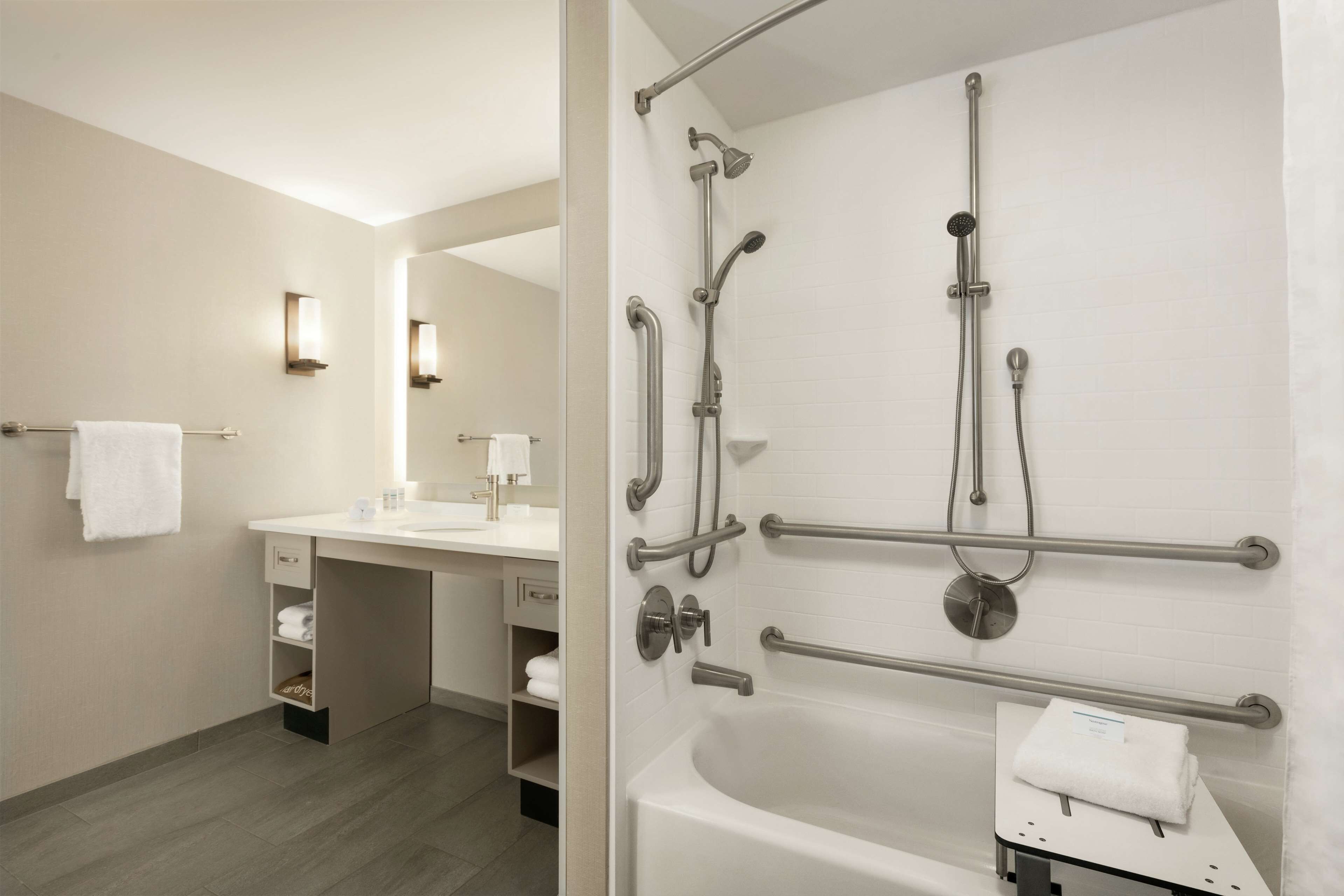 Un baño moderno con lavamanos blanco, espejo y bañera. Incluye barras de apoyo y cabezal de ducha de mano.