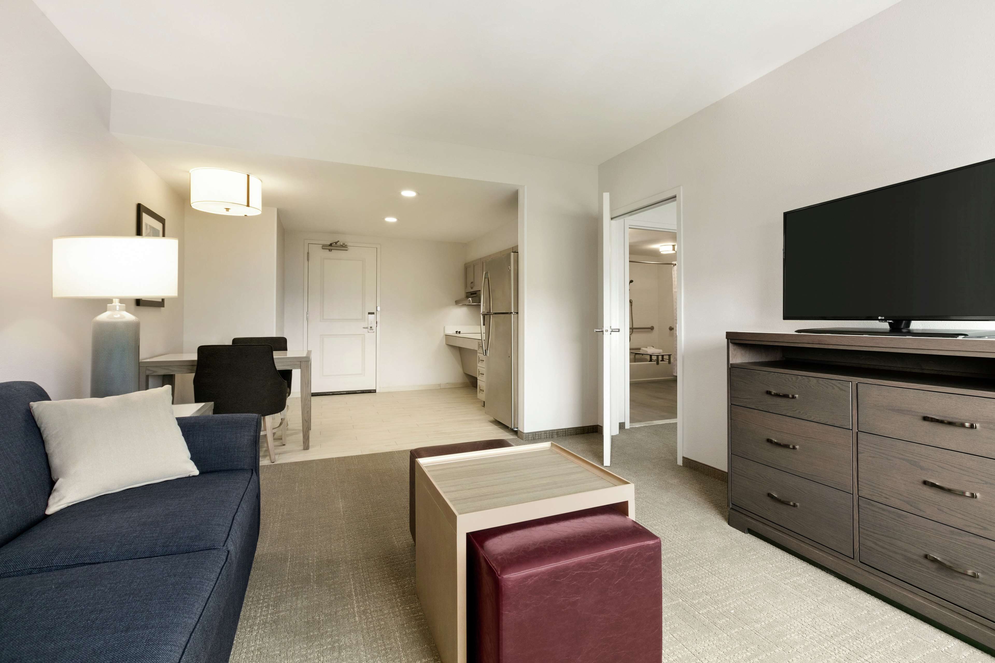 A modern hotel room with a sofa, a TV, a dining area, and a kitchen in the background.