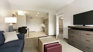 A modern hotel room with a sofa, a TV, a dining area, and a kitchen in the background.
