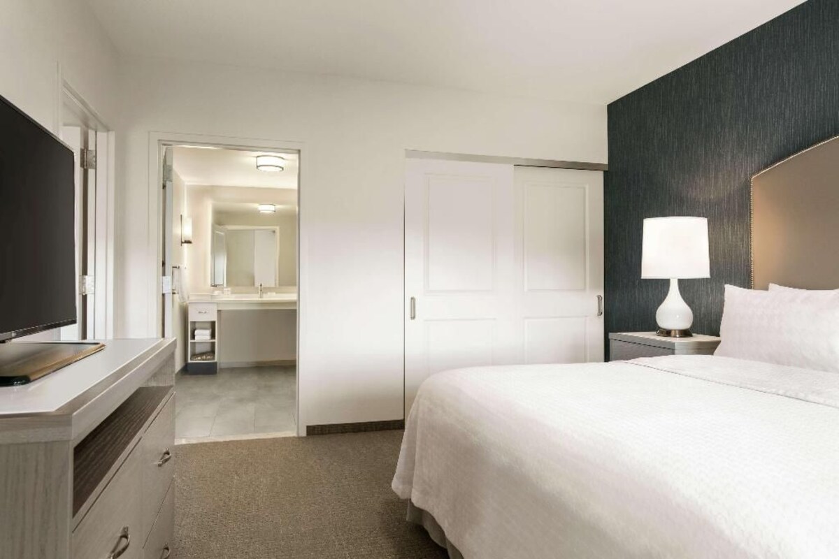 Une chambre d’hôtel équipée d’un lit, d’un bureau, d’une télévision, d’un miroir et d’une porte donnant accès à une salle de bain.